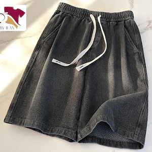 Puede incluir: Pantalones cortos grises oscuros con cintura elástica y cordón blanco. Los pantalones cortos tienen bolsillos laterales y un corte holgado. La tela parece ser mezclilla texturizada. El logo "CLOTHES BY ILAN" está en la esquina superior izquierda.