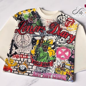 Puede incluir: Camiseta de manga corta color crema con estampado gráfico colorido. El estampado presenta las palabras "Lovers Diary" en rojo, junto con varias ilustraciones, incluyendo la Estatua de la Libertad, símbolos de la paz y calaveras. La camiseta tiene cuello redondo.