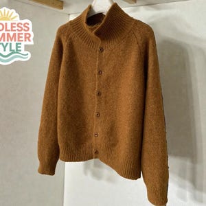 以下が含まれることがあります： ハイネックとボタン留めの茶色のカーディガン。セーターは柔らかく質感のある素材でできており、白いハンガーに吊るされています。「ENDLESS SUMMER STYLE」の文字が左上にあります。