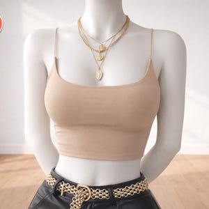 Puede incluir: Un top corto beige con tirantes finos se muestra en un maniquí. Un cinturón de cadena dorada y collares dorados en capas con colgantes completan el look. El maniquí está sobre un fondo blanco.