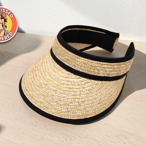 Può includere: Visiera di paglia con finiture e fascia nere. La visiera è realizzata in materiale di paglia intrecciata e ha una tesa curva. L'immagine include anche un adesivo circolare con la scritta "Sun-Kissed & Covered".