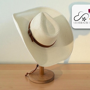 May include: A cream-colored cowboy hat with a brown leather band and a wooden hat stand. The hat has a wide brim and a classic cowboy hat shape. The hat stand is made of light-colored wood. The logo "CLOTHES BY ILAN" is in the upper right corner.