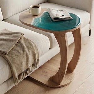 May include: A stylish side table with a wooden base and a tabletop featuring a teal resin design resembling ocean waves. A coffee cup and a magazine are placed on the table. The table is next to a white sofa.