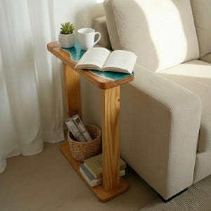 Könnte beinhalten: Beistelltisch aus Holz mit hellblauem Epoxidharz-Oberteil. Der Tisch hat ein unteres Regal mit Büchern und einem Weidenkorb. Eine weiße Tasse, eine kleine Topfpflanze und ein offenes Buch liegen auf der Tischoberfläche.