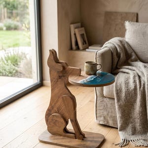 Puede incluir: Una mesa auxiliar de madera con forma de perro sentado, que presenta un tablero circular con una incrustación de resina azul que imita las olas del mar. Una taza de cerámica descansa sobre la superficie de la mesa, apoyada en una base rectangular plana.