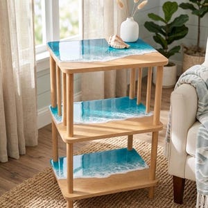 May include: A three-tiered wooden side table with a coastal theme. The shelves feature a blue and white resin design resembling ocean waves. A white vase with dried flowers and a seashell decorate the top shelf. The table is set on a woven jute rug.