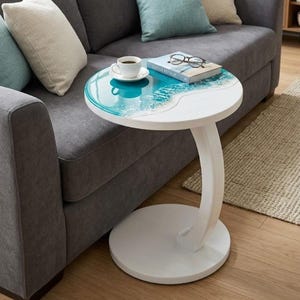 May include: A round white side table with a curved pedestal base. The tabletop features a decorative resin inlay depicting a turquoise ocean wave with white foam detailing. A white coffee cup, a book titled Seaside Retreat, and glasses sit on top.