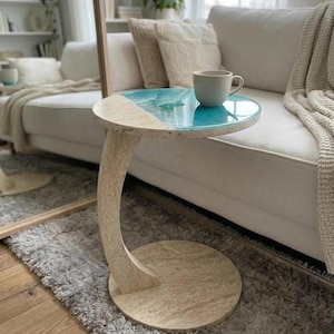 May include: A C-shaped side table with a circular base and top made of beige speckled stone. The tabletop features a decorative inlay of translucent blue resin that mimics the appearance of ocean waves along one curved edge, topped with a mug.