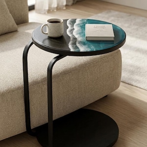 May include: A black side table with a round top featuring a blue and white ocean wave design. A white mug with coffee and a book titled "Coastal Musings" rest on the table. The table has a black, curved metal frame.
