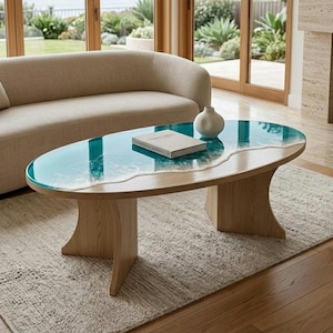 May include: An oval coffee table with a light wood base and a tabletop featuring a blue resin inlay that mimics the appearance of ocean waves with white foam detailing. A white ceramic vase and a closed book sit on the table surface.