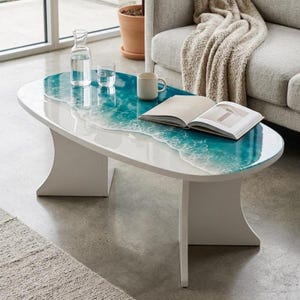 May include: An oval coffee table with a white base and a tabletop featuring a blue and white resin ocean wave design. The table holds a clear glass carafe, a small glass, a white ceramic mug, and an open book with white pages and text.