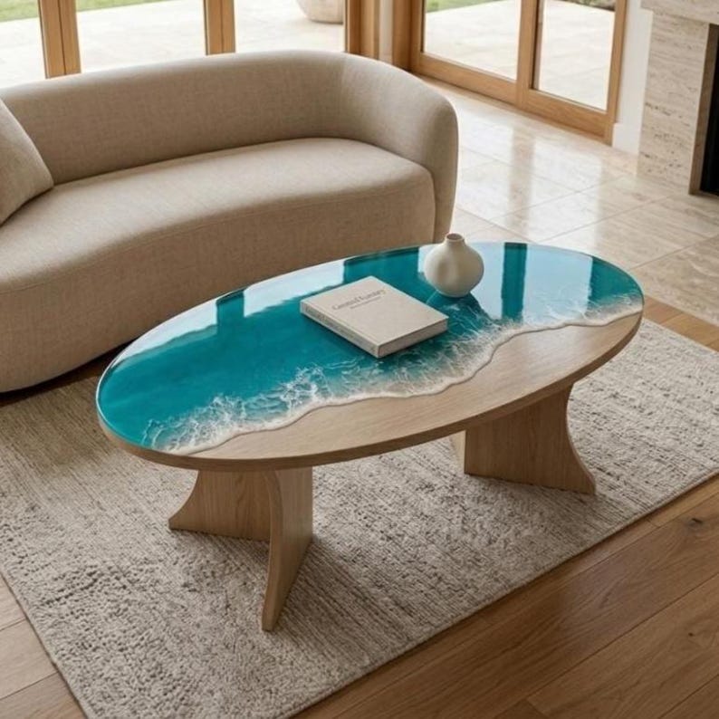 May include: An oval coffee table with a light wood base and a tabletop featuring a teal resin inlay that mimics ocean waves with white foam detailing. A white ceramic vase and a closed book are placed on the smooth, glossy surface.