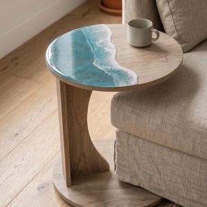 May include: A round side table with a wooden base and a tabletop featuring a blue and white resin design resembling ocean waves. A small, gray mug sits on the wooden portion of the table. The table is next to a beige sofa.