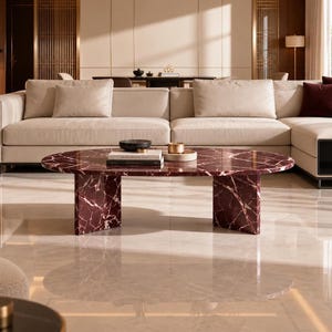May include: An oval coffee table crafted from deep burgundy marble with prominent white veining, supported by two thick, rectangular slab legs. The tabletop holds a small black bowl, a stack of books, and a circular tray with a metallic object.