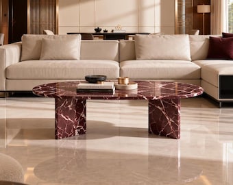 May include: An oval coffee table crafted from deep burgundy marble with prominent white veining, supported by two thick, rectangular slab legs. The tabletop holds a small black bowl, a stack of books, and a circular tray with a metallic object.
