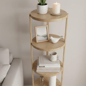 Puede incluir: Una estantería redonda de cuatro niveles de madera clara. El estante superior contiene una planta en maceta, una vela encendida y un pequeño marco de fotos de madera. Los estantes inferiores muestran un pájaro de cerámica, libros, una taza y botellas decorativas.