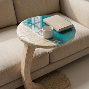 Pode incluir: Uma mesa lateral redonda com um design único. O tampo da mesa apresenta um design de resina azul e branco inspirado no oceano. Uma caneca branca e um livro estão sobre a mesa. A mesa é suportada por uma base de pedra bege curva.