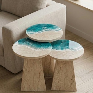 Può includere: Tre tavolini con tema costiero. Ogni tavolo ha un piano rotondo con un design di spiaggia e oceano nei toni del blu, bianco e beige. Le basi sono di colore beige chiaro e hanno una forma trapezoidale.