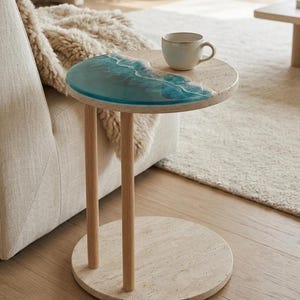 May include: A circular side table with a distinctive design. The tabletop incorporates a section of blue resin art, mimicking ocean waves. It has three wooden legs and a circular base. A small, light-coloured mug rests on the table.