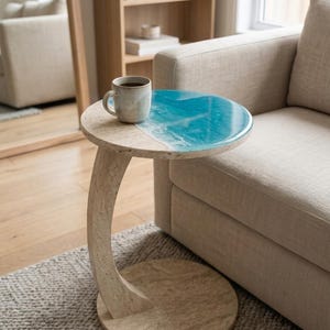 May include: A C-shaped side table with a beige travertine stone base and curved support, featuring a circular top with a smooth, glossy blue and white resin inlay that mimics ocean waves, topped with a speckled ceramic mug.