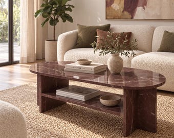 May include: An oval coffee table made of dark reddish-brown marble with white veining. The table features a smooth top surface and a lower shelf supported by two vertical slab legs. The surface holds a small bowl and a book titled KIN F O L K.