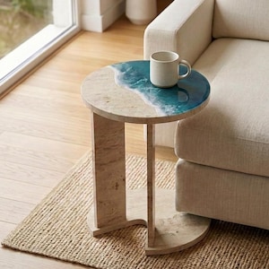 May include: A round side table made of beige travertine stone with a glossy blue resin inlay that mimics the appearance of ocean waves crashing onto a sandy beach. A speckled off-white ceramic mug sits on the table surface.