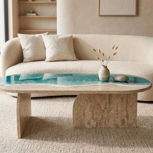 May include: An oval coffee table featuring a beige stone base and a tabletop with a smooth, light-colored stone surface on one side and a glossy, teal-colored resin inlay that mimics the appearance of ocean waves with white foam detailing.