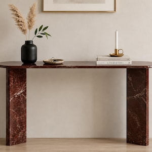 May include: A dark red marble console table with a smooth, elongated oval top and two thick, rectangular slab legs. The surface features natural white veining throughout the stone, providing a polished finish to this furniture piece.