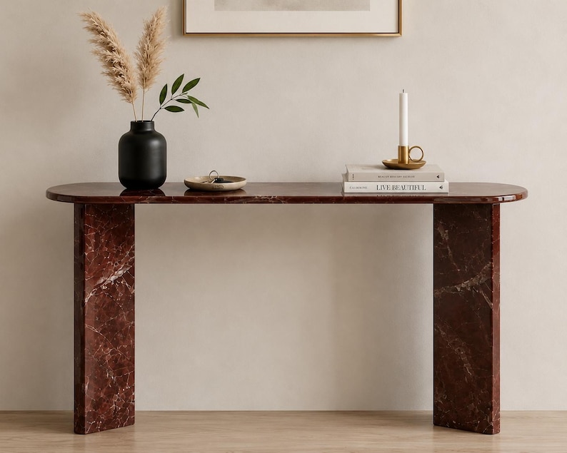 May include: A dark red marble console table with a smooth, elongated oval top and two thick, rectangular slab legs. The surface features natural white veining throughout the stone, providing a polished finish to this furniture piece.