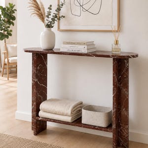 May include: A rectangular console table made of dark red marble with white veining, featuring a flat top surface and a lower shelf. The table has two vertical slab legs and a smooth finish, providing a sturdy structure for displaying various home decor items.