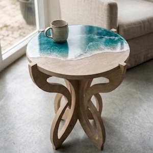 Op de afbeelding: Een ronde bijzettafel met een epoxyhars blad met oceaanthema. Het tafelblad heeft een blauw en wit golfpatroon. De tafel heeft een licht houten onderstel met een kenmerkend, gebogen pootontwerp. Een keramische mok staat op de tafel.