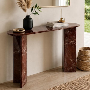 May include: A dark red marble console table with a smooth, polished surface and two rectangular legs. The table features a long, rounded top and a distinct veined pattern throughout the stone. It is styled with a black vase, books, and a candle.