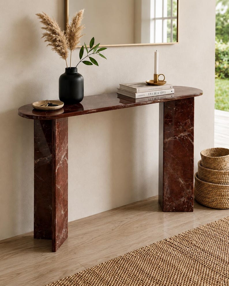 May include: A dark red marble console table with a smooth, polished surface and two rectangular legs. The table features a long, rounded top and a distinct veined pattern throughout the stone. It is styled with a black vase, books, and a candle.
