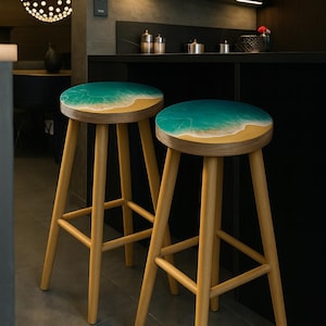 May include: Two wooden bar stools with a beach-themed design. The round seats feature a turquoise ocean scene with a sandy shore. The stools have light-colored wooden legs and a footrest.