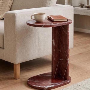 May include: A C-shaped side table crafted from deep red marble with prominent white veining. The table features a smooth, polished surface with a rounded top and a matching rounded base connected by a vertical slab, supporting a ceramic mug and a notebook.