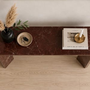May include: A dark red marble console table with a smooth, rounded-edge top and two sturdy legs. On the surface sits a black vase with dried pampas grass, a small beige dish containing keys, and a stack of books topped with a gold candle holder.