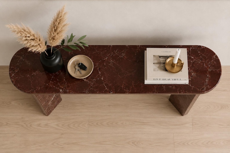 May include: A dark red marble console table with a smooth, rounded-edge top and two sturdy legs. On the surface sits a black vase with dried pampas grass, a small beige dish containing keys, and a stack of books topped with a gold candle holder.