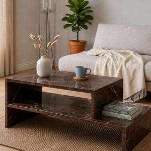 May include: A rectangular coffee table with a dark brown marble finish featuring a tiered design with an open storage shelf. The table surface holds a white ceramic vase with dried stems, a light blue mug on a coaster, and two stacked books.