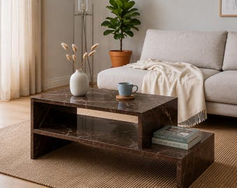 May include: A rectangular coffee table with a dark brown marble finish featuring a tiered design with an open storage shelf. The table surface holds a white ceramic vase with dried stems, a light blue mug on a coaster, and two stacked books.