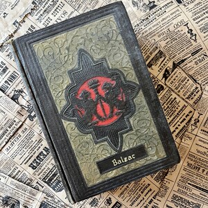 May include: A vintage hardback book with a dark green and black cover. The cover features a gold embossed design with two mythical creatures flanking a central symbol. The book's title, "Balzac", is printed in gold lettering on the front cover.