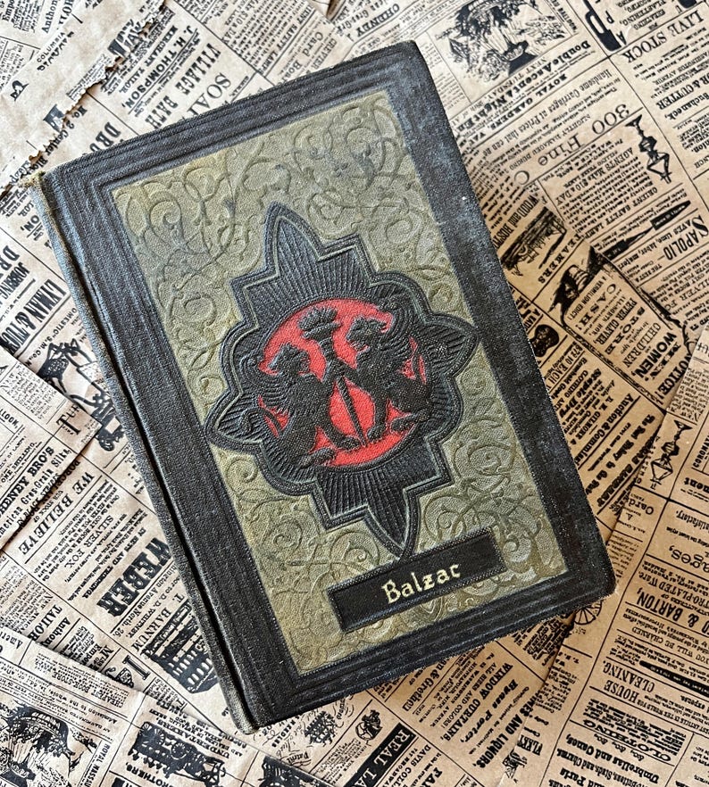 May include: A vintage hardback book with a dark green and black cover. The cover features a gold embossed design with two mythical creatures flanking a central symbol. The book's title, "Balzac", is printed in gold lettering on the front cover.