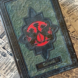 May include: A vintage, dark green leather-bound book with a gold embossed design featuring two griffins flanking a torch. The title "Tolstoi" is embossed on the front cover.