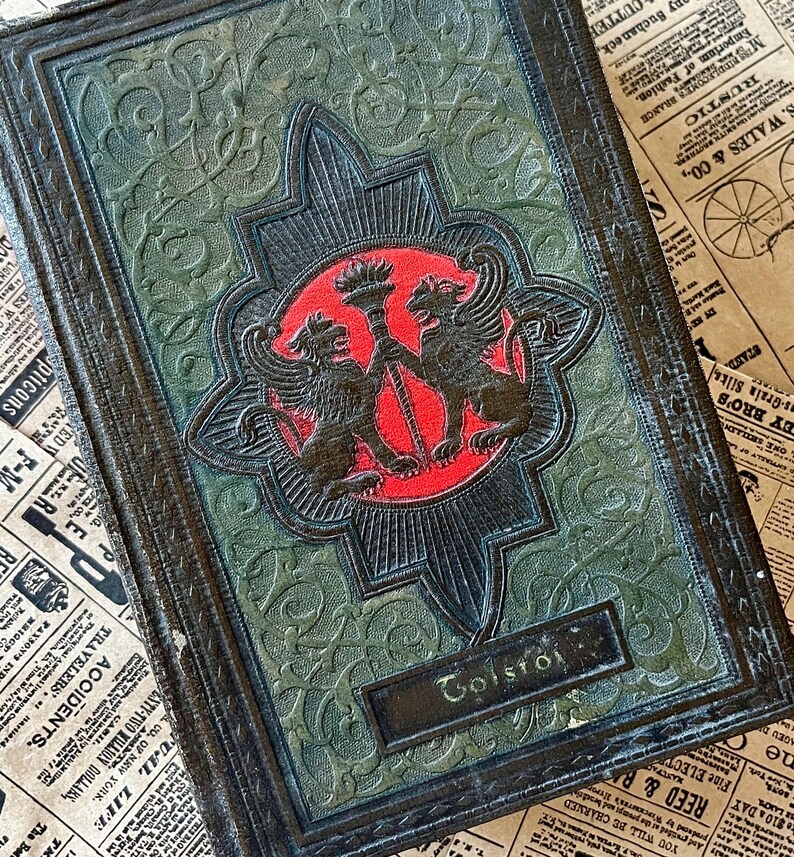 May include: A vintage, dark green leather-bound book with a gold embossed design featuring two griffins flanking a torch. The title "Tolstoi" is embossed on the front cover.