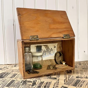 May include: A wooden box with a hinged lid, open to reveal vintage items. Inside are glass jars, a key, a padlock, and a small photograph. The box is made of light brown wood and has brass hinges. The background is a newspaper print.