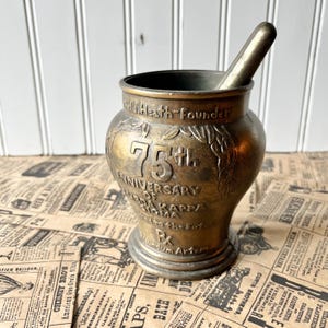 May include: A vintage brass mortar and pestle. The mortar is inscribed with "75th Anniversary of Kappa Sigma" and "W. Heath Founder." The pestle rests inside the mortar. The background includes newspaper clippings and a white-painted wood surface.
