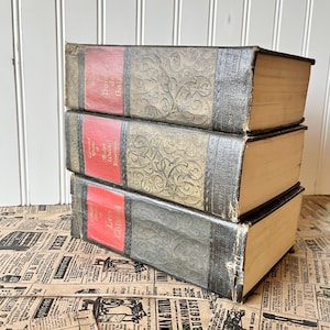 May include: A stack of three vintage hardback books with ornate gold designs on the covers. The books have red spines with gold lettering. The books are on a brown and white patterned surface.