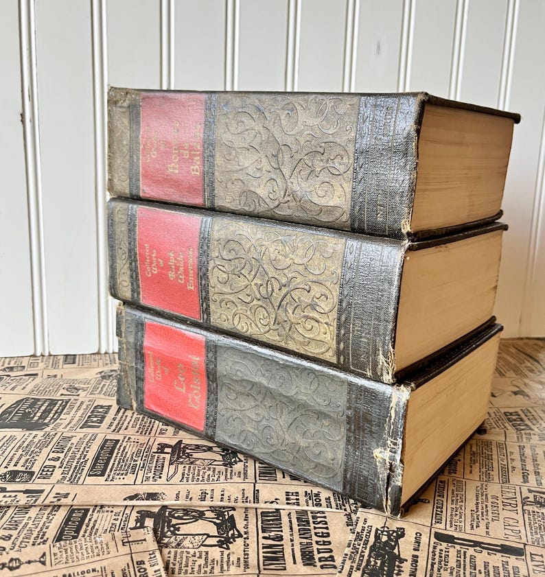 May include: A stack of three vintage hardback books with ornate gold designs on the covers. The books have red spines with gold lettering. The books are on a brown and white patterned surface.