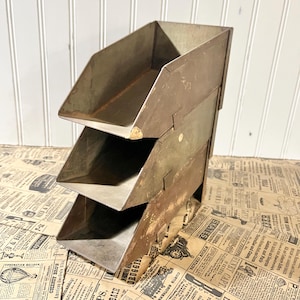 May include: A vintage metal organizer with three angled trays. The metal has a weathered, aged appearance with visible rust and wear. The trays are designed to hold papers or small items. The organizer is a practical and decorative storage solution.