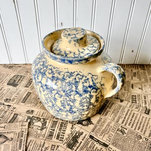 May include: A vintage stoneware crock with a lid, featuring a cream-colored base and a blue, speckled pattern. The crock has a handle and a matching lid with a knob. The background includes a newspaper and a white-paneled wall.