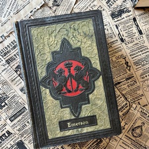 May include: A vintage hardback book with a dark green and gold cover. The cover features an embossed design with two mythical creatures holding a torch. The title "Emerson" is printed in gold lettering on the front cover.